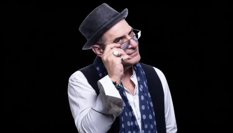 Francesco Baccini con cappello grigio, occhiali scuri e foulard blu a pois, ritratto su sfondo nero