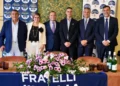 Sei persone in posa dietro un tavolo con fiori e lo striscione di Fratelli d’Italia, nella sala della conferenza a Scicli