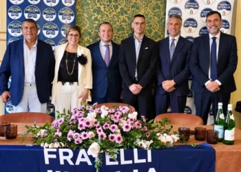 Sei persone in posa dietro un tavolo con fiori e lo striscione di Fratelli d’Italia, nella sala della conferenza a Scicli