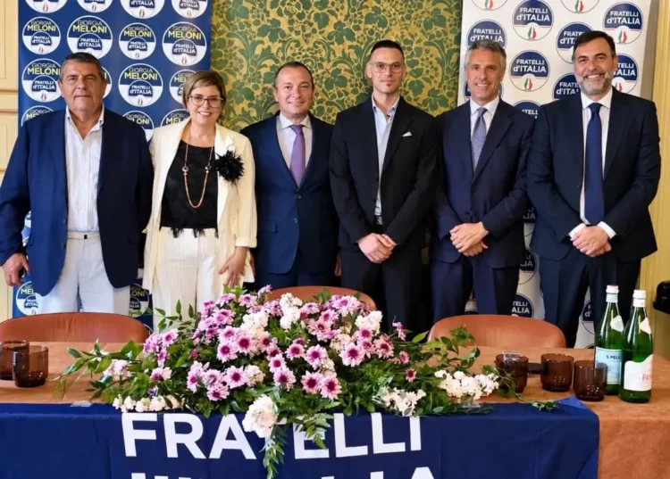Sei persone in posa dietro un tavolo con fiori e lo striscione di Fratelli d’Italia, nella sala della conferenza a Scicli