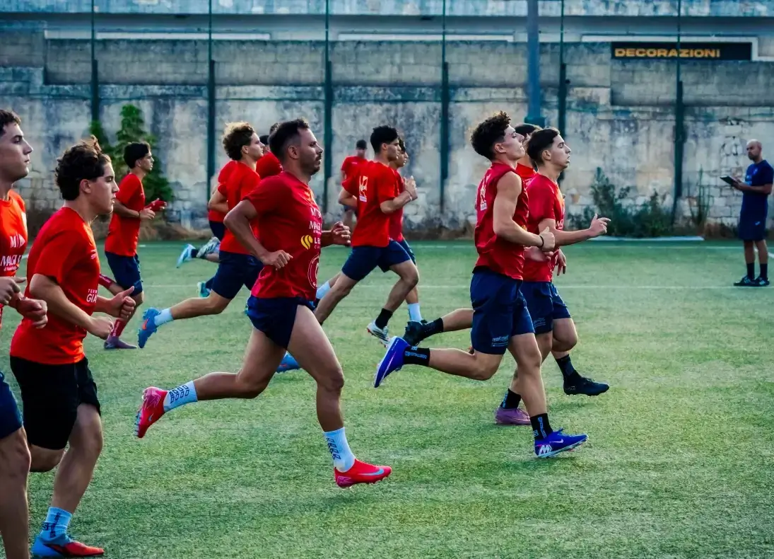 Calciatori del Frigintini Calcio in maglia rossa e pantaloncini blu corrono in allenamento su un campo in erba sintetica