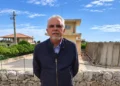 Ignazio Abbate in giacca blu davanti a un muretto a secco, con una torre e una vasca in cemento sullo sfondo