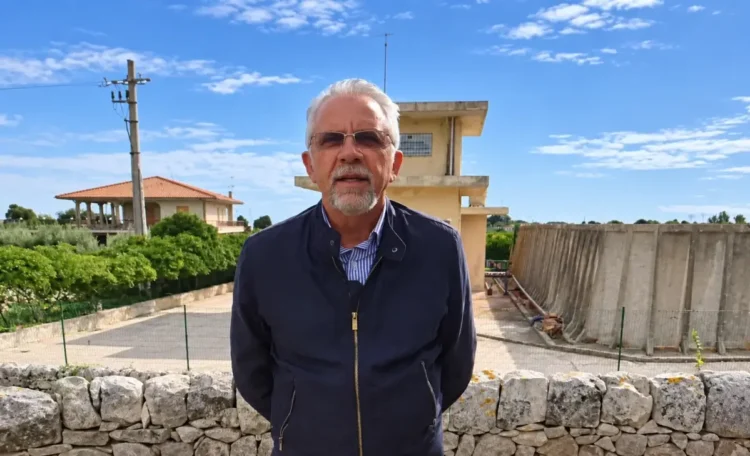 Ignazio Abbate in giacca blu davanti a un muretto a secco, con una torre e una vasca in cemento sullo sfondo