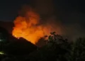 Incendio notturno: alte fiamme arancioni e una colonna di fumo su un pendio collinare, alberi in controluce in primo piano