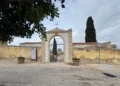 Ingresso ad arco del cimitero di Santa Croce Camerina con cancello in ferro, mura gialle, un cipresso e il piazzale davanti