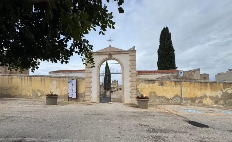 Ingresso ad arco del cimitero di Santa Croce Camerina con cancello in ferro, mura gialle, un cipresso e il piazzale davanti