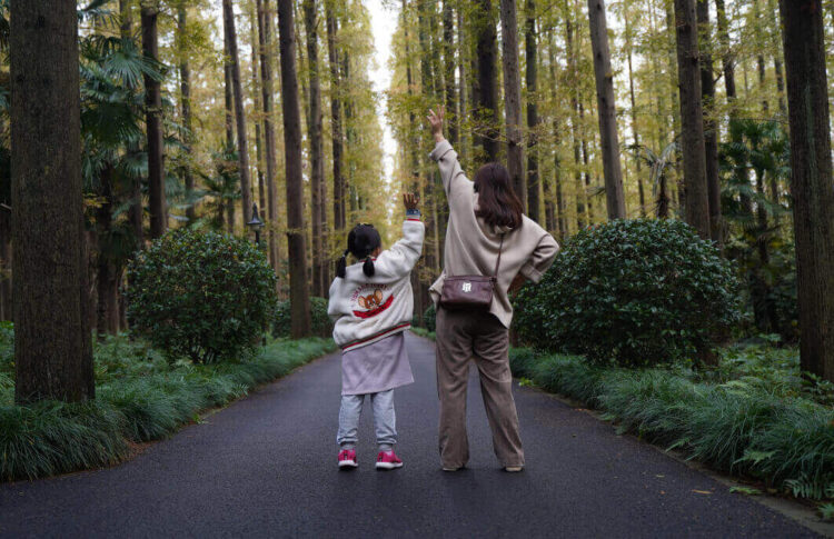 Bonus mamme 2026: una madre e una bambina di spalle alzano il braccio lungo un viale alberato in un bosco
