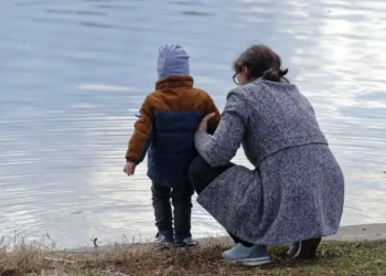 Una madre accovacciata di spalle tiene il figlio piccolo in riva a un lago, immagine per il bonus mamme 2026