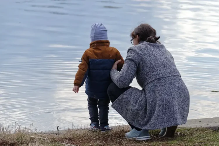 Una madre accovacciata di spalle tiene il figlio piccolo in riva a un lago, immagine per il bonus mamme 2026