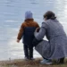 Una madre accovacciata di spalle tiene il figlio piccolo in riva a un lago, immagine per il bonus mamme 2026