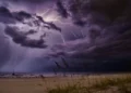 Fulmini tra nubi violacee e pioggia sul mare in tempesta, con una barca a vela e l’erba delle dune piegata dal vento