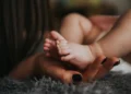 Mano di una mamma con le unghie smaltate di scuro che sorregge i piedini di un neonato sdraiato su un tappeto grigio