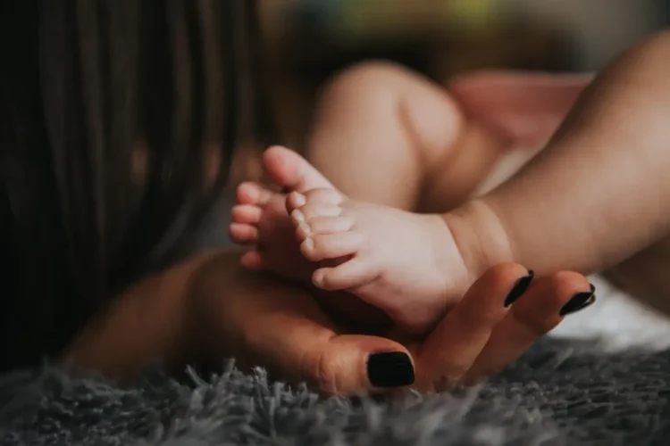 Mano di una mamma con le unghie smaltate di scuro che sorregge i piedini di un neonato sdraiato su un tappeto grigio