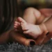 Mano di una mamma con le unghie smaltate di scuro che sorregge i piedini di un neonato sdraiato su un tappeto grigio