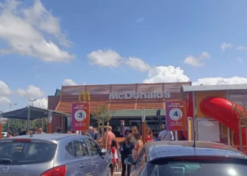 Ingresso di un ristorante McDonald’s con clienti in coda, auto parcheggiate in primo piano e area giochi con scivolo rosso