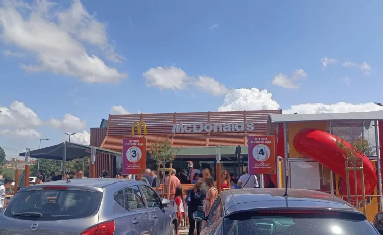 Ingresso di un ristorante McDonald’s con clienti in coda, auto parcheggiate in primo piano e area giochi con scivolo rosso