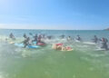 Surfisti in mare con le braccia alzate attorno a una tavola coperta di fiori, nell’omaggio a Felix Castro Lopez