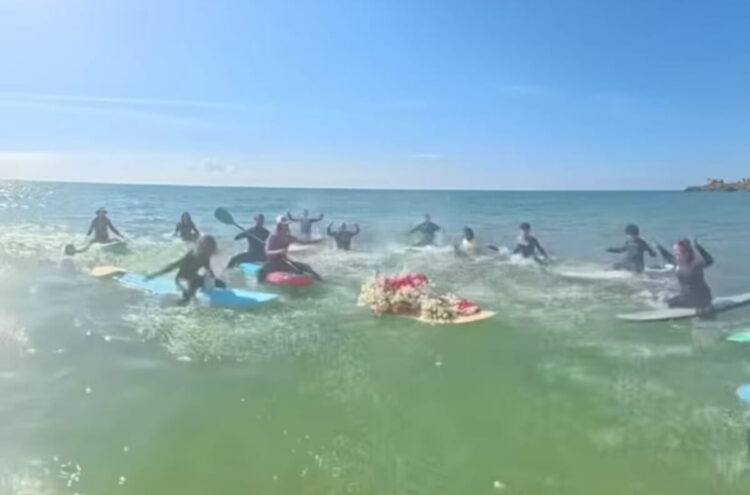 Surfisti in mare con le braccia alzate attorno a una tavola coperta di fiori, nell’omaggio a Felix Castro Lopez