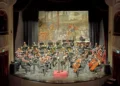 Orchestra sul palco del Teatro Garibaldi di Modica, con il direttore su un podio rosso e un fondale dipinto con carri romani