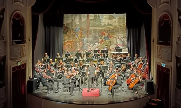 Orchestra sul palco del Teatro Garibaldi di Modica, con il direttore su un podio rosso e un fondale dipinto con carri romani