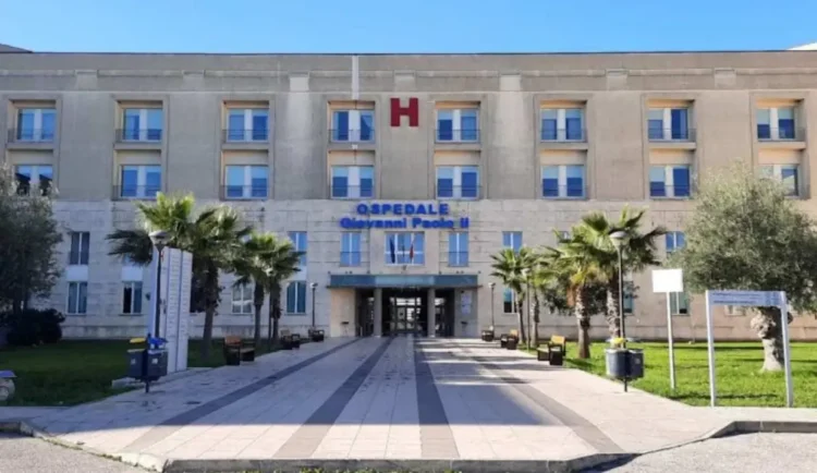 Ingresso dell’ospedale Giovanni Paolo II di Ragusa, con la scritta blu sulla facciata, la H rossa e un viale tra le palme