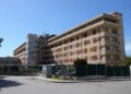 Ospedale Maggiore di Modica: edificio a più piani con tapparelle gialle e rosse, recinzione verde e auto parcheggiate