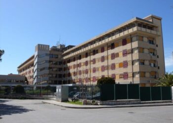 Ospedale Maggiore di Modica: edificio a più piani con tapparelle gialle e rosse, recinzione verde e auto parcheggiate