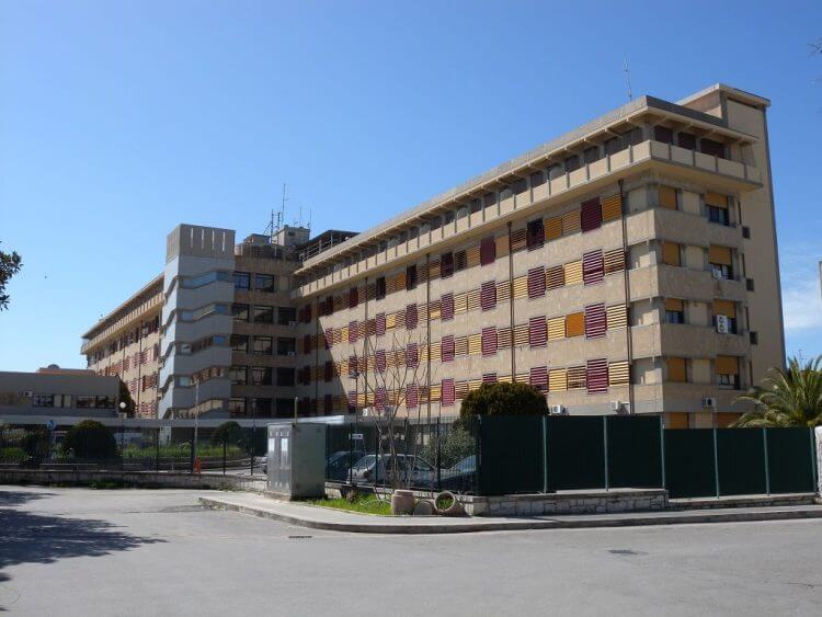 Ospedale Maggiore di Modica: edificio a più piani con tapparelle gialle e rosse, recinzione verde e auto parcheggiate