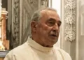 Padre Salvatore Dinatale con l’abito talare chiaro accanto a un altare decorato con stucchi bianchi e candelieri dorati