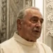 Padre Salvatore Dinatale con l’abito talare chiaro accanto a un altare decorato con stucchi bianchi e candelieri dorati
