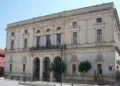 Facciata in pietra chiara del Palazzo di Città di Ragusa, con tre arcate, balcone centrale e bandiere, sotto un cielo sereno