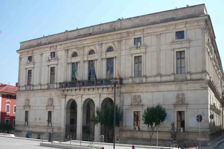 Facciata in pietra chiara del Palazzo di Città di Ragusa, con tre arcate, balcone centrale e bandiere, sotto un cielo sereno