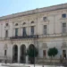 Facciata in pietra chiara del Palazzo di Città di Ragusa, con tre arcate, balcone centrale e bandiere, sotto un cielo sereno