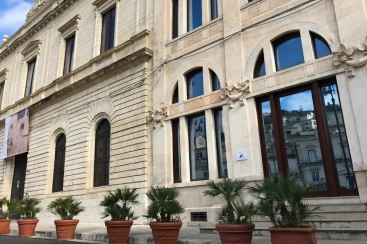 Facciata in pietra di un palazzo del centro storico di Modica, con finestre ad arco, vetrate e palme in vaso all’ingresso