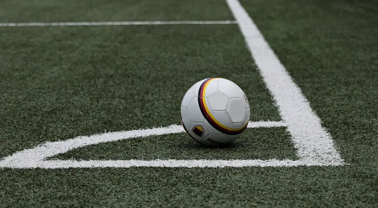 Pallone da calcio bianco con strisce nere, rosse e gialle fermo vicino alla bandierina del calcio d’angolo su un campo in erba sintetica