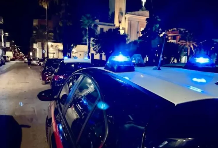 Auto dei carabinieri con lampeggianti blu accesi di notte in una strada del centro di Vittoria, con una chiesa illuminata sullo sfondo