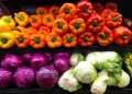 Peperoni gialli, arancioni e rossi esposti su un ripiano, sotto cavoli rossi, cavoli verza e lattughe