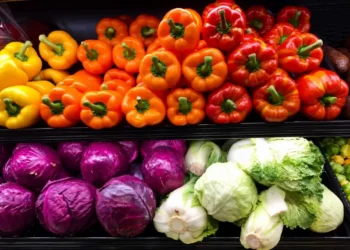 Peperoni gialli, arancioni e rossi esposti su un ripiano, sotto cavoli rossi, cavoli verza e lattughe