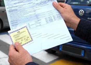 Mani di un uomo con il modulo di una polizza auto e il contrassegno assicurativo giallo, davanti a un’auto parcheggiata