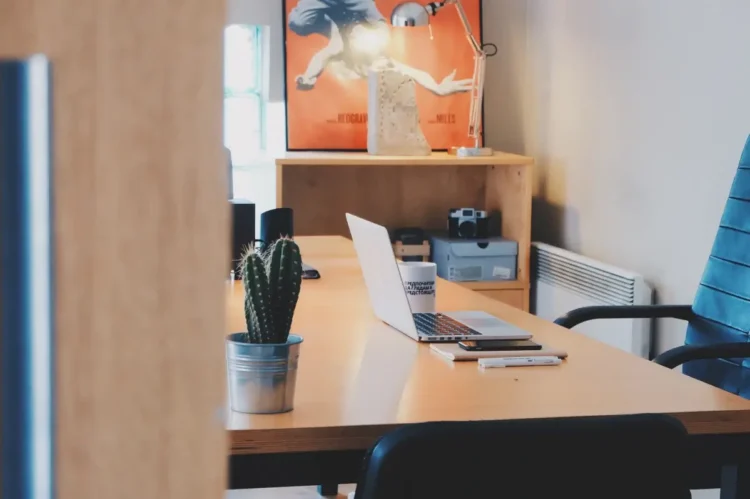 Scrivania in legno con computer portatile, tazza, smartphone e cactus in vaso in un ufficio con sedia blu e scaffale