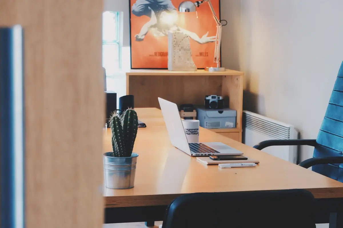 Scrivania in legno con computer portatile, tazza, smartphone e cactus in vaso in un ufficio con sedia blu e scaffale