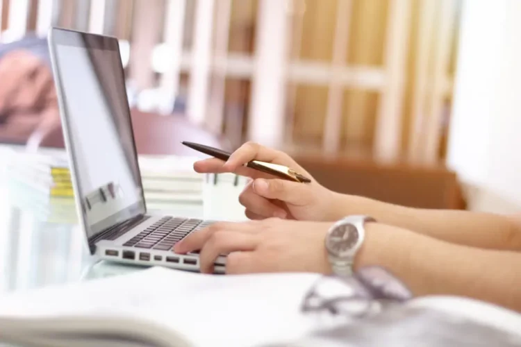 Mani al lavoro su un portatile con una penna, accanto a libri e fogli: scrivania di uno studio, tema incentivi alle imprese