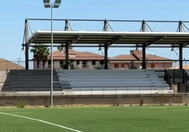 Tribuna coperta con struttura in acciaio e gradinate grigie davanti a un campo da calcio in erba sintetica a Ragusa, impianto sportivo