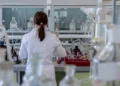 Ricercatrice in camice bianco di spalle al banco di un laboratorio con vetreria, foto per la ricerca su artrosi ed enzima
