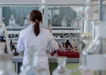 Ricercatrice in camice bianco di spalle al banco di un laboratorio con vetreria, foto per la ricerca su artrosi ed enzima
