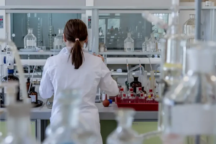 Ricercatrice in camice bianco di spalle al banco di un laboratorio con vetreria, foto per la ricerca su artrosi ed enzima