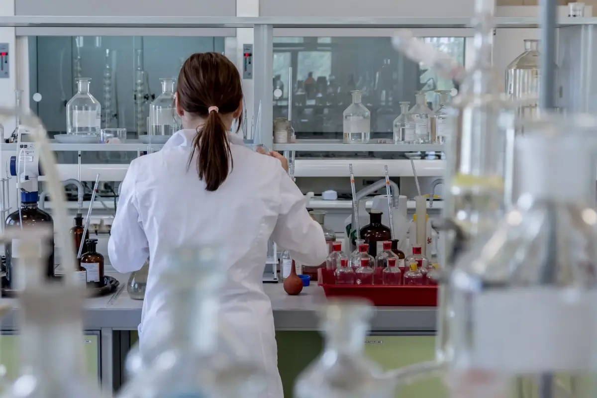 Ricercatrice in camice bianco di spalle al banco di un laboratorio con vetreria, foto per la ricerca su artrosi ed enzima