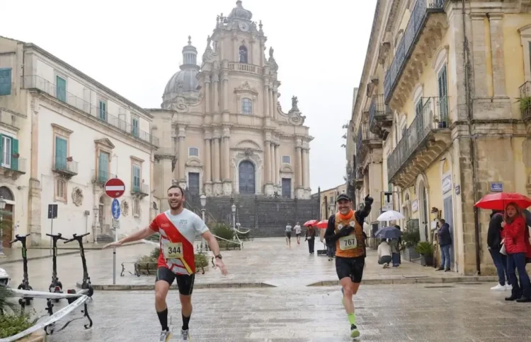 Due runner con pettorale corrono la maratona di Ragusa in piazza Duomo a Ibla, sotto la pioggia, con San Giorgio sullo sfondo