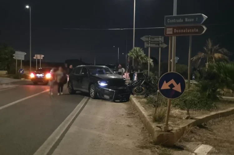 Incidente auto moto a Marina di Ragusa: Audi scura con il paraurti divelto e la moto a terra accanto all’aiuola, di sera