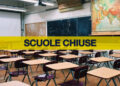 Aula scolastica vuota con banchi, sedie, lavagna e carta geografica, attraversata da una fascia gialla con la scritta «Scuole chiuse»